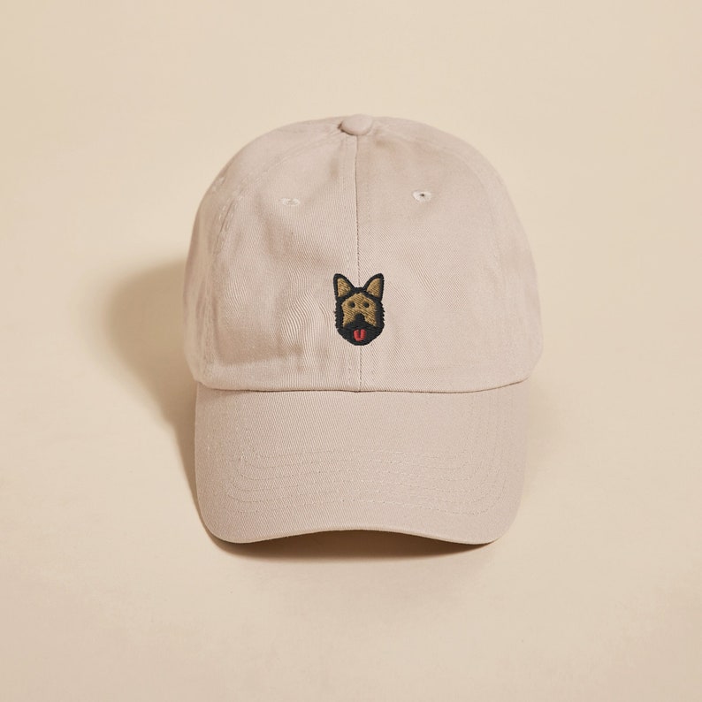 May include: A beige baseball cap with an embroidered brown and black dog face with a red tongue.