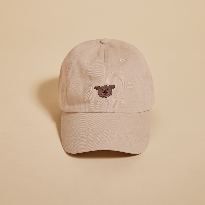 K&ouml;nnte beinhalten: Eine beige Baseballkappe mit einem braunen gestickten Hundefoto auf der Vorderseite.