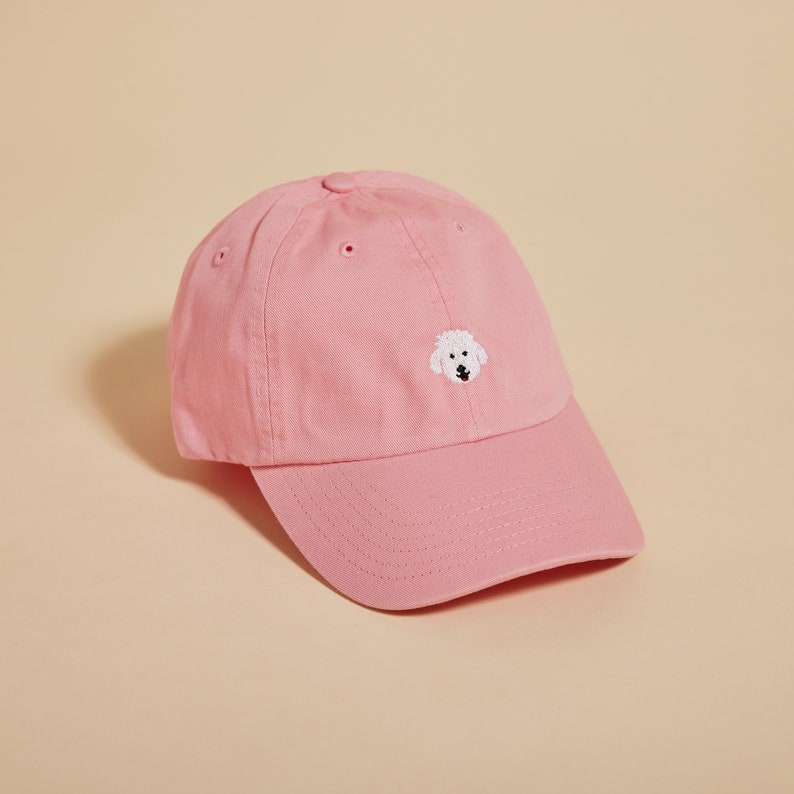May include: A pink baseball cap with a white embroidered dog face on the front.