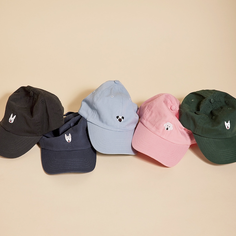 May include: Five baseball caps with different embroidered dog faces. The caps are black, light blue, pink, dark blue, and dark green.