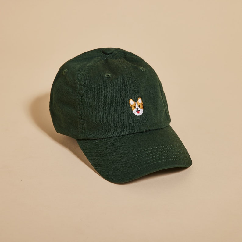 K&ouml;nnte beinhalten: Eine dunkelgr&uuml;ne Baseballkappe mit einem gestickten Bild eines Corgi-Hundes auf der Vorderseite.