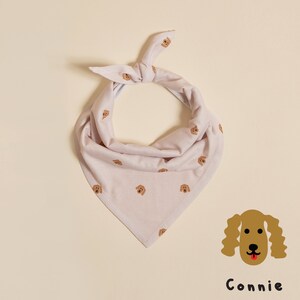 May include: A light pink bandana with a repeating pattern of brown dog faces. The bandana is tied in a knot at the top and has a triangular shape. The text "Connie" is printed below a brown dog face.