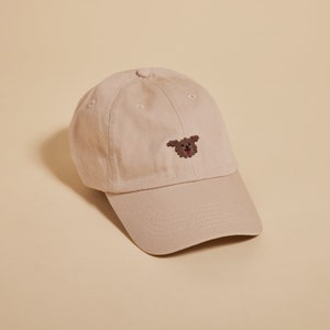 K&ouml;nnte beinhalten: Eine beige Baseballkappe mit einem braunen gestickten Hundefoto auf der Vorderseite.