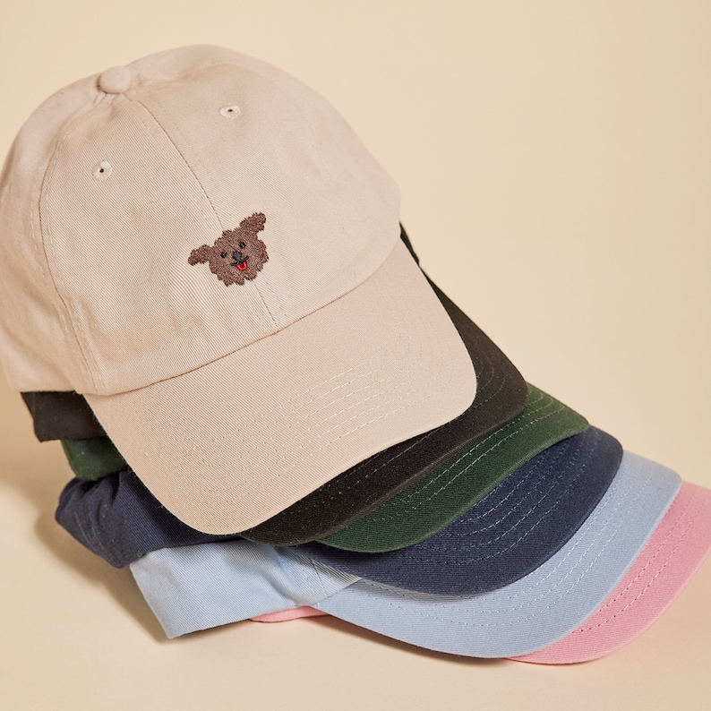 K&ouml;nnte beinhalten: Ein Stapel von sechs Baseballkappen in verschiedenen Farben, darunter beige, schwarz, gr&uuml;n, marineblau, hellblau und rosa. Die oberste Kappe zeigt einen gestickten braunen Hund mit roter Zunge.