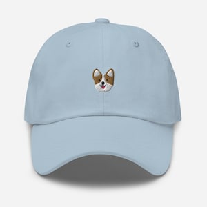 K&ouml;nnte beinhalten: Eine hellblaue Baseballkappe mit einem gestickten Bild eines braunen und wei&szlig;en Hundes mit rosa Zunge.