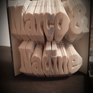 Könnte beinhalten: Eine Buchkunstskulptur mit den Namen "Marcel & Nadine", die in die Seiten gefaltet sind. Das Buch ist geöffnet und die Seiten sind zu einem 3D-Design gefaltet.
