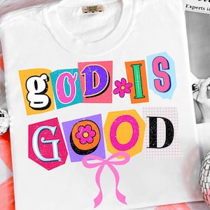 May include: White t-shirt with the phrase "GOD IS GOOD" in colorful, cut-out style letters. The design includes a pink bow and flower accents. The shirt is a casual, everyday wear item.