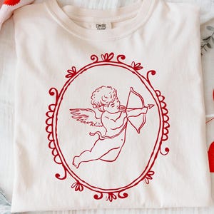 Pu&ograve; includere: T-shirt color crema con un'illustrazione rossa di Cupido che punta il suo arco e le frecce all'interno di una cornice ovale. La maglietta &egrave; piegata su una superficie bianca, con decorazioni di San Valentino sullo sfondo.