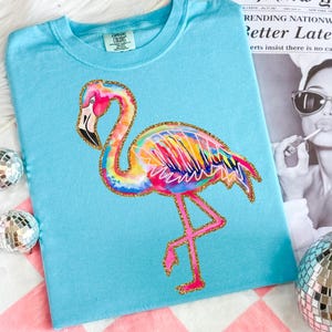 May include: A light blue t-shirt featuring a colorful flamingo design with gold glitter accents. The flamingo has a rainbow-colored body and pink legs. The shirt is on a pink and white checkered surface, with a newspaper clipping and disco balls.