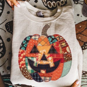May include: A light grey sweatshirt with a patchwork pumpkin design. The pumpkin is made of various fabric patterns in orange, red, green, and blue. The pumpkin has a smiling jack-o'-lantern face and a brown stem. The sweatshirt is folded.