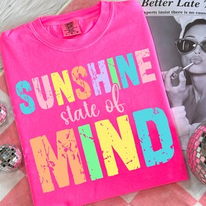 May include: A bright pink Comfort Colors t-shirt with the words "SUNSHINE state of MIND" in colorful, distressed lettering. The shirt is laid flat on a red and white checkered surface, with a disco ball and a black and white image in the background.