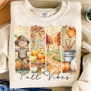 Puede incluir: Sudadera crema con un gráfico temático de otoño. El diseño presenta un espantapájaros, calabazas, un camino y las palabras "Fall Vibes" en una fuente cursiva. La paleta de colores incluye naranja, marrón y verde.