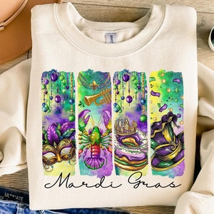 May include: A cream colored sweatshirt with a watercolor design of Mardi Gras symbols. The symbols include a mask, a lobster, a crown, and a hat. The text "Mardi Gras" is written in black below the symbols.