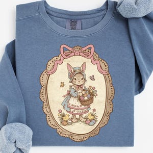 May include: A blue sweatshirt featuring an Easter-themed illustration. The design includes a bunny holding a basket of eggs, set within an oval frame with a pink bow. The sweatshirt has a crew neck and long sleeves.