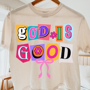 May include: A beige t-shirt with the words "GOD IS GOOD" in colorful, cut-out style letters. The design includes a pink bow and flower accents. The shirt is hanging on a wooden hanger.