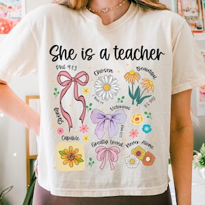 Op de afbeelding: Cr&egrave;mekleurig T-shirt met de tekst "She is a teacher" in zwart schrift. Het shirt heeft bloemen- en lintafbeeldingen met woorden als "Chosen", "Beautiful" en "Capable".
