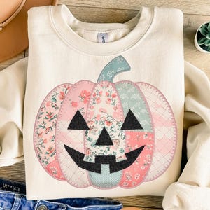 May include: Cream-coloured sweatshirt featuring a patchwork pumpkin design. The pumpkin has a smiling face with black triangle eyes and mouth. The pumpkin is made of pink, blue, and white floral patterned fabric. A fun and festive autumn-themed top.