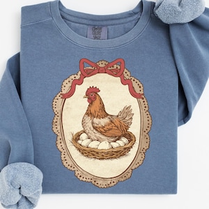 Peut inclure: Un sweat-shirt bleu avec une illustration de poule. La poule est marron et blanche, assise sur un nid d'œufs, dans un cadre ovale avec un nœud rouge. Le sweat-shirt a un col rond et des manches longues avec un poignet doux et flou.