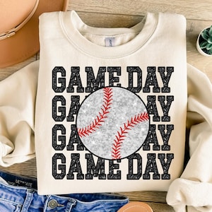 以下が含まれることがあります： クリーム色のスウェットシャツに、黒い文字で「GAME DAY」が4回繰り返され、中央にはキラキラ光る銀色の野球ボールが描かれています。