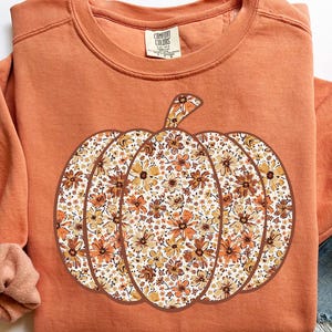 May include: Orange Comfort Colors sweatshirt with a large pumpkin design. The pumpkin is filled with a floral pattern in shades of orange, brown, and cream. The sweatshirt has a crew neck and long sleeves, perfect for fall.