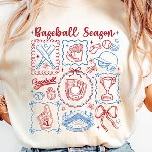 May include: Cream-colored t-shirt with a baseball-themed graphic. The design includes baseball bats, a glove, a stadium, and the text "Baseball Season." The graphic is in red and blue, with a decorative border.