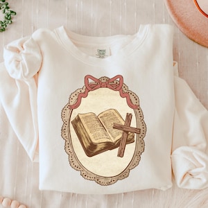 May include: Cream-colored sweatshirt featuring a framed illustration of an open book and a wooden cross. The illustration is in a vintage style, with a pink bow at the top of the frame. The sweatshirt has long sleeves and a crew neck.