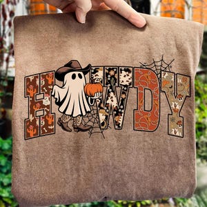 May include: A brown sweatshirt featuring the word "HOWDY" in a Western-style font. The letters are decorated with cacti, cow print, and skulls. A ghost in a cowboy hat and boots holds a pumpkin, with a spider web design.
