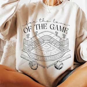 Puede incluir: Sudadera color crema con un gráfico en blanco y negro de un estadio de béisbol, béisbol, guante y bates. El texto "for the love OF THE GAME" está encima del gráfico. Un estilo casual y cómodo.