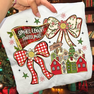 Puede incluir: Funda de almohada blanca con temática navideña con un diseño festivo. Presenta una pancarta a cuadros con el texto "Small Town Christmas", dos lazos decorativos y una escena de pueblo con casas y un árbol de Navidad.