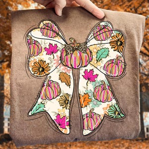 Puede incluir: Camiseta marrón con un gran diseño de lazo con temática otoñal. El lazo está decorado con calabazas, flores y hojas en tonos naranja, rosa y verde. El lazo tiene un centro con estampado de leopardo.