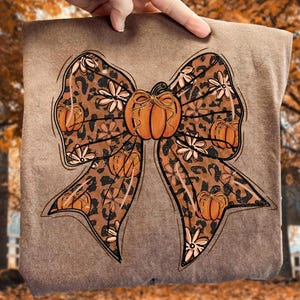 May include: A brown sweatshirt featuring a large bow design with a leopard print pattern. The bow is adorned with orange pumpkins and white floral accents, perfect for fall. The design is held up by a hand.