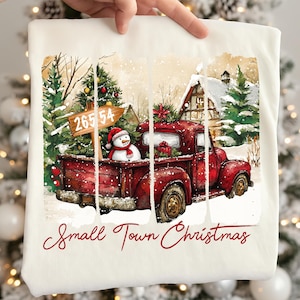 Może przedstawiać: Biała bluza z ilustracją czerwonej ciężarówki w stylu vintage w zimowym krajobrazie. Bałwan i prezenty znajdują się w ciężarówce. Tekst "Small Town Christmas" jest napisany czerwoną czcionką.