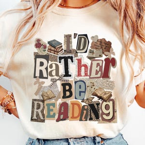 Peut inclure: T-shirt crème avec un motif de collage affichant le texte « I'd Rather Be Reading ». Le design comprend des images de livres, de papillons et d'éléments floraux. Les lettres sont dans différentes polices et couleurs, créant un effet superposé.