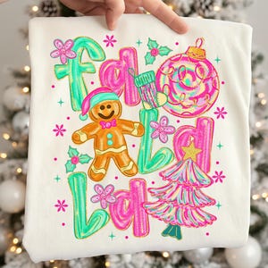 Puede incluir: Camiseta de color crema con un diseño navideño colorido. El diseño incluye las palabras "Fa La La La" en letras de burbujas rosas y verdes, un hombre de jengibre, un árbol de Navidad y adornos.