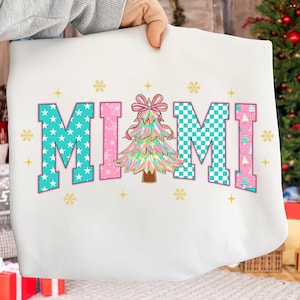 Puede incluir: Sudadera blanca con la palabra "MIAMI" en letras de bloque turquesa y rosa. Las "M" son turquesas con estrellas blancas y a cuadros, la "A" es rosa y las "I" son turquesas con diseños de árboles de Navidad. Un colorido árbol de Navidad está en el centro.