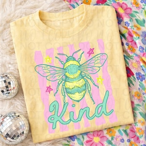 以下が含まれることがあります： 明るい黄色のTシャツ。カラフルな蜂のデザインと「Kind」の文字が特徴です。蜂は黄色、青、緑で、ピンクのストライプと黄色の星が背景です。Tシャツは折りたたまれています。