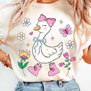 May include: A white t-shirt featuring a cartoon illustration of a goose wearing pink boots and a pink bow. The goose is surrounded by flowers and a butterfly.