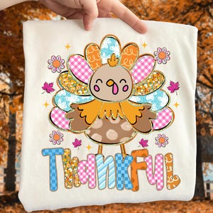 May include: A cream-colored t-shirt featuring a colorful turkey design with the word "Thankful" in a playful font. The turkey has a brown body, orange feathers, and a cheerful expression. Floral and leaf accents surround the turkey.