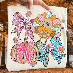May include: Cream-colored t-shirt featuring a Thanksgiving-themed design. The design includes a cartoon turkey, a pumpkin with a pink bow, and two bows with floral and plaid patterns. Additional floral accents and a fall leaf complete the design.