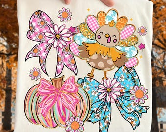 May include: Cream-colored t-shirt featuring a Thanksgiving-themed design. The design includes a cartoon turkey, a pumpkin with a pink bow, and two bows with floral and plaid patterns. Additional floral accents and a fall leaf complete the design.