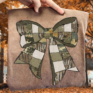 Può includere: Un tessuto marrone con un grande motivo a fiocco. Il fiocco presenta un motivo patchwork con sezioni mimetizzate e a righe bianche. Il fiocco è nei toni del verde, marrone e beige. Lo sfondo è sfocato, con foglie autunnali.