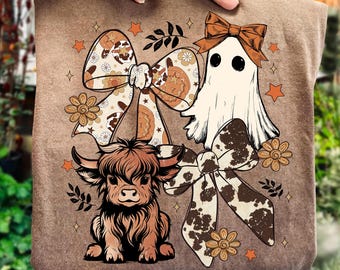 May include: A brown t-shirt with a quirky design featuring a ghost, a Highland cow, and decorative bows. The design incorporates floral and star accents, using a palette of brown, orange, and cream.