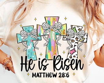 May include: A white t-shirt with a graphic of three colorful crosses with bows. The text "He is Risen" and "Matthew 28:6" is printed below the crosses.