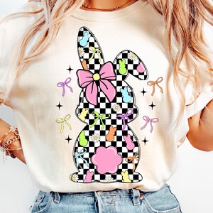 May include: A white t-shirt with a checkered bunny silhouette design. The bunny is decorated with pastel-colored bows, flowers, and candy.