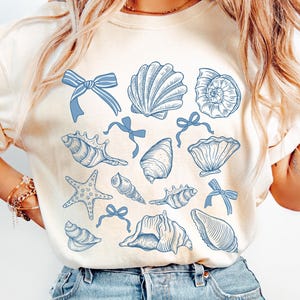 May include: A white t-shirt featuring a blue seashell design. The design includes various seashells, starfish, and bows.