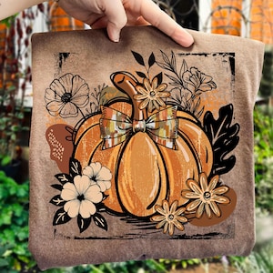 May include: A brown tote bag with an orange pumpkin design, complete with a plaid bow and floral elements. The artwork has a vintage feel, ideal for autumn. The bag is held up by a hand.