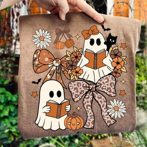 May include: Brown t-shirt featuring a whimsical Halloween design. Two ghost figures are reading books, surrounded by bows, flowers, pumpkins, stars, and a black cat. The design incorporates orange, brown, and white colors, creating a festive and playful aesthetic.