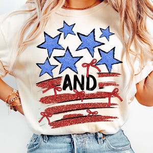 Pode incluir: Uma camiseta branca com um design de bandeira americana vermelha, branca e azul. A bandeira tem estrelas azuis e la&ccedil;os vermelhos. A palavra "AND" est&aacute; escrita em letras pretas na bandeira.