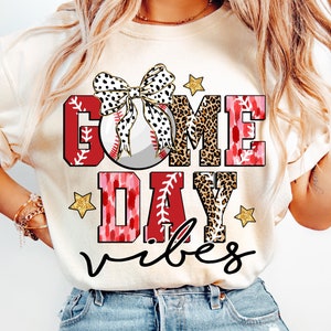 以下が含まれることがあります： クリーム色のTシャツに、赤、黒、白の文字で「Game Day Vibes」と書いてあります。文字は、ヒョウ柄、ピンクと赤のタイダイ、黒いリボンが付いた白い野球ボールで飾られています。