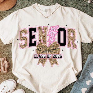 Puede incluir: Camiseta color crema con la palabra "SENIOR" en negro y estampado de leopardo, un rayo rosa y un lazo con estampado de leopardo. El texto "CLASS OF 2026" está debajo del lazo. La camiseta está rodeada de accesorios.